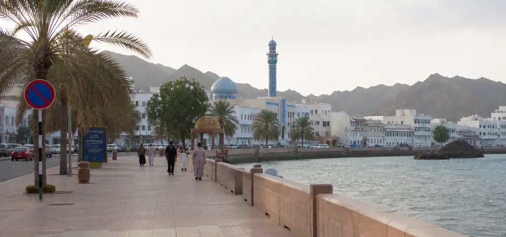 Expat Walking Along A Muscat Beach Promenade With Sea And Mountains In The Background