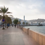 Expat Walking Along A Muscat Beach Promenade With Sea And Mountains In The Background