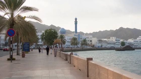Expat Walking Along A Muscat Beach Promenade With Sea And Mountains In The Background
